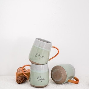 May include: Three ceramic mugs in a stack, one lying on its side. The mugs are light green with a gray rim and orange handle. The word "Oma" is written on two of the mugs. A small wooden box with coasters and a pinecone are in the background.