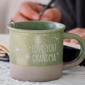 Könnte beinhalten: Grüne Keramiktasse mit weißem Henkel. Die Tasse hat eine hellgrüne Glasur und eine weiße Aufschrift mit dem Text "Love You Grandma". Die Tasse ist mit einem weißen Löwenzahnmuster verziert.