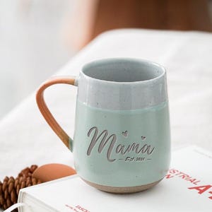 Puede incluir: Una taza de cerámica con cuerpo verde claro y borde gris. La palabra "Mama" está grabada en la parte delantera, con "Est. 2021" debajo. El asa es de color marrón cálido. La taza está sobre un libro blanco.