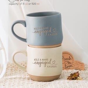 Puede incluir: Dos tazas de cerámica con un esmalte beige y azul. Las tazas están apiladas una encima de la otra. La taza superior es azul y la taza inferior es beige. Ambas tazas tienen el texto "KILI & MAKE engaged 6.4.2025" en una fuente cursiva.