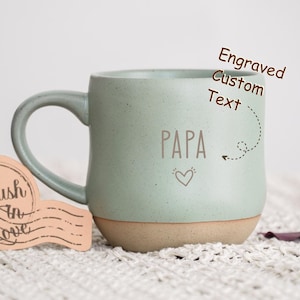 May include: A light green ceramic mug with a brown rim and handle. The mug has the word "PAPA" written on it in brown lettering with a small heart symbol below. The mug is sitting on a white fluffy surface. The text "Engraved Custom Text" is written in brown lettering above the mug.