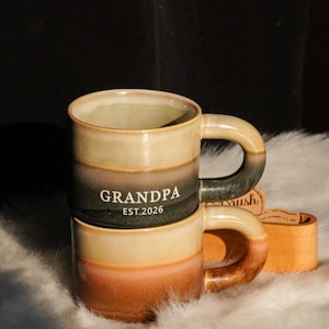 Peut inclure: Deux mugs en céramique empilés avec un dégradé de beige, noir et orange. Le mug du dessus porte l'inscription "GRANDPA EST. 2026" en lettres blanches. Les mugs ont de grandes anses incurvées et sont posés sur une surface blanche et duveteuse.