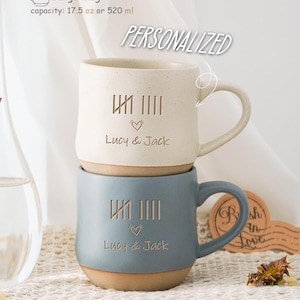 Op de afbeelding: Twee gepersonaliseerde keramische mokken met een lichtblauwe en witte glazuur. De mokken hebben een ontwerp van Romeinse cijfers en een hart met de tekst "Lucy & Jack". De mokken zijn 17,5 ounce of 520 milliliter.
