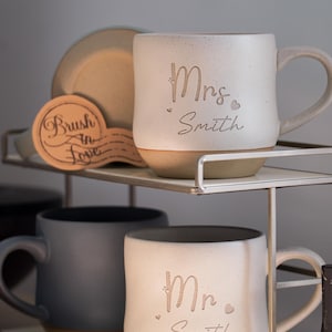 May include: Ceramic mugs with "Mrs. Smith" and "Mr. Smith" etched on them. The mugs are off-white with a tan base and handle. A wooden coaster with the text "Brush in Love" is also visible.