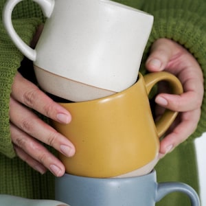 May include: Three ceramic mugs stacked on top of each other. The top mug is white, the middle mug is yellow, and the bottom mug is blue.