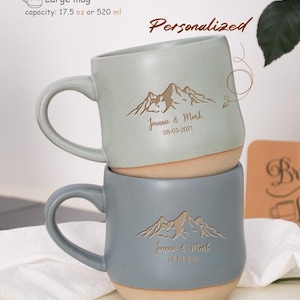 以下が含まれることがあります： 山をモチーフにしたデザインと「Joannie & Mark」というテキスト、そして「08-03-2021」という日付が書かれた、パーソナライズされたセラミック製のマグカップ2個。マグカップは薄い青色とベージュで、容量は520mlです。