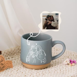 Könnte beinhalten: Eine blaue Keramiktasse mit hellbraunem Boden. Die Tasse zeigt eine weiße Strichzeichnung eines sich umarmenden Paares, das ein Selfie macht. Ein kleines Foto des Paares befindet sich über der Tasse. Die Tasse steht auf einer weißen Häkeloberfläche.