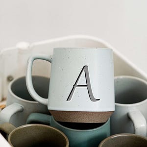 May include: A light blue ceramic mug with the letter "A" in a dark gray font. The mug has a brown base and a handle. Several other mugs in various shades of blue and green are visible in the background.