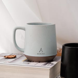 May include: A light blue ceramic mug with a brown base and handle. The mug has a speckled texture and features the letter "A" and the date "03.06.2025". A black mug is visible to the right.