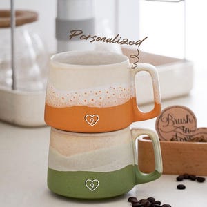 Puede incluir: Dos tazas de cerámica apiladas con un diseño en capas. La taza superior es naranja y crema, la inferior es verde y crema. Cada taza tiene un corazón con el número 9. La palabra "Personalizado" está escrita encima de las tazas.