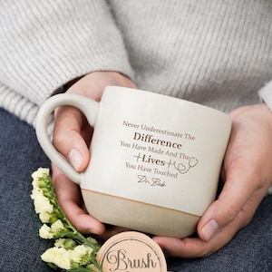 Puede incluir: Una taza de cerámica color crema con base beige, sostenida por una persona. La taza presenta el texto "Never Underestimate The Difference You Have Made And The Lives You Have Touched Dr. Bob" y un gráfico de estetoscopio. También hay una pequeña ramita de flores amarillas y una etiqueta que dice "Brush In Love".