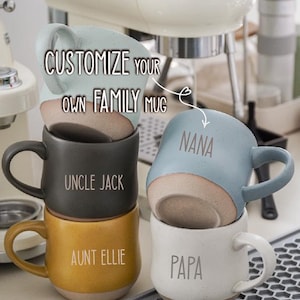 Può includere: Pila di tazze in ceramica in vari colori, tra cui blu, nero, giallo e bianco. Ogni tazza ha una base chiara e un manico. Le tazze sono personalizzate con nomi di famiglia come "NANA", "UNCLE JACK", "AUNT ELLIE" e "PAPA". È visibile anche il testo "CUSTOMIZE YOUR OWN FAMILY MUG".