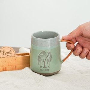 May include: A ceramic mug with a light green body, gray rim, and a brown handle. The mug features a weeping willow tree design with the initials "M A" and the date "02.11.2016".