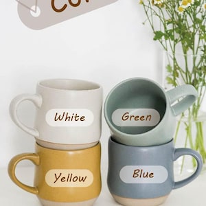 May include: Four ceramic mugs stacked on top of each other. The mugs are white, green, yellow, and blue. Each mug has a label with the color of the mug written on it.