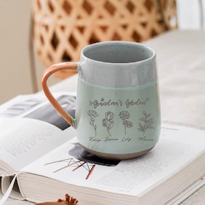 Puede incluir: Taza de cerámica con esmalte verde claro y gris, con el texto "Grandma's Garden" e ilustraciones florales. El asa es de color naranja cálido. La taza está sobre un libro abierto.