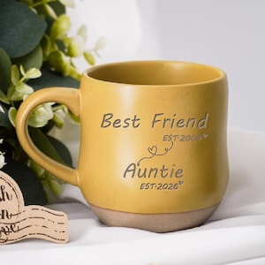 May include: Mustard yellow ceramic mug with a curved handle. The mug has the words "Best Friend EST 2004" and "Auntie EST 2026" etched on the front. The mug is sitting on a white surface with greenery and a decorative wooden tag.
