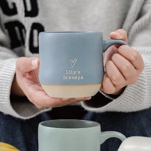 May include: A blue ceramic mug with a beige base, featuring the text "Lily's Grandpa" and a heart symbol. The mug has a handle and is held by a person. The mug is a light blue colour.