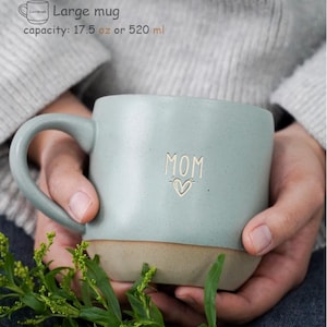 May include: A large, light blue ceramic mug with a beige base. The mug features the word "MOM" and a heart design on the front. The mug has a capacity of 520 ml.