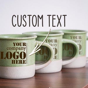 Peut inclure: Trois tasses en céramique vertes avec des bords blancs. Les tasses ont un motif de texte brun qui dit "Your company logo here" sur une tasse et "Your company logo here" sur les deux autres tasses.