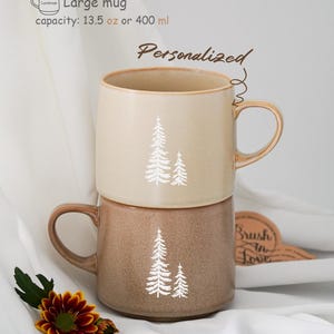 Puede incluir: Dos tazas de cerámica con un esmalte marrón y beige. Las tazas presentan una silueta blanca de un diseño de pino. Las tazas están apiladas una encima de la otra. La taza superior tiene el texto "Personalizado" escrito en ella. El texto "Taza grande capacidad: 13,5 oz o 400 ml" está escrito encima de las tazas.