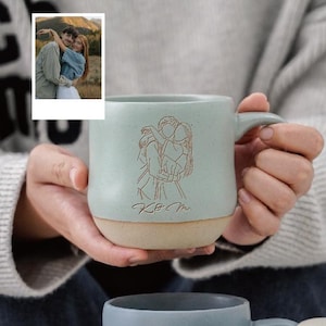 Może przedstawiać: Jasnoniebieski ceramiczny kubek z beżową podstawą, z grawerowaną ilustracją obejmującej się pary. Kubek ma uchwyt, a inicjały "K&M" są wygrawerowane pod ilustracją. Małe zdjęcie jest w tle.