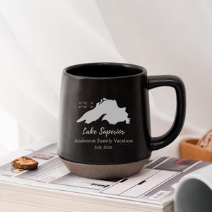 Può includere: Una tazza in ceramica nera con una sagoma grigio chiaro del Lago Superiore, le coordinate 47°42' N 87°30' W e il testo Lake Superior, Anderson Family Vacation, July 2026. La tazza presenta una parte superiore nera liscia e una base marrone ruvida.