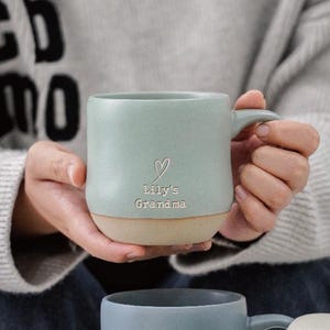 以下が含まれることがあります： ベージュのベースとハート、そして「Lily's Grandma」の文字が入った、薄い緑色のセラミックマグカップ。 マグカップにはカーブしたハンドルがあり、人が持っています。 マグカップはマグカップのコレクションの一部です。