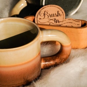 May include: A ceramic mug with a gradient of colors, transitioning from beige to orange-brown. The handle is a darker brown. A wooden holder with the text "Brush In" is visible, along with other ceramic items.