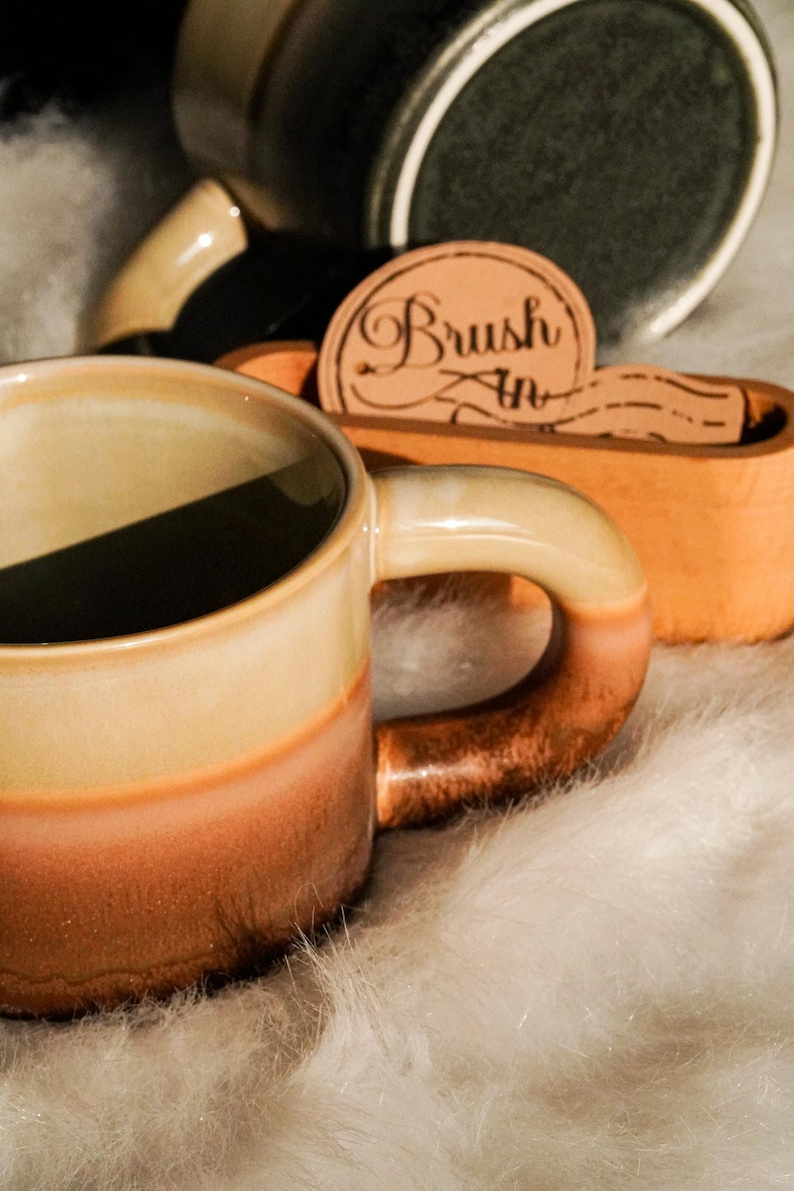 May include: A ceramic mug with a gradient of colors, transitioning from beige to orange-brown. The handle is a darker brown. A wooden holder with the text "Brush In" is visible, along with other ceramic items.