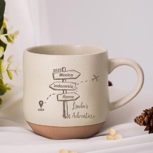 May include: Cream-colored ceramic mug with a brown base, featuring a travel-themed design. The mug has a handle and displays directional signs pointing to Mexico, Indonesia, and Rome, with the text "Linda's Adventure."