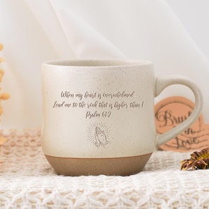 Puede incluir: Taza de cerámica color crema con base marrón y asa curvada. La taza presenta el texto "When my heart is overwhelmed, Lead me to the rock that is higher than I, Psalm 61:2" y una ilustración de manos rezando.