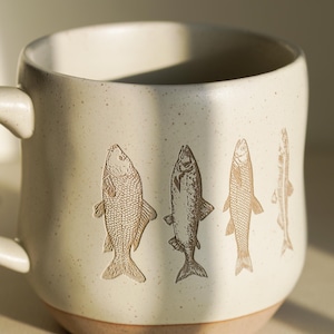 May include: A white ceramic mug with a brown rim and four fish designs on the side. The fish are in a line and are facing to the right. The mug is on a white surface.