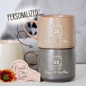 May include: Two stacked ceramic mugs, one tan and one gray, with a personalized calendar design. The tan mug has the text "25 March 1997 Javier & Angellina". The gray mug has the text "25 March 1997 Jessica & Angellina". A third mug is visible.