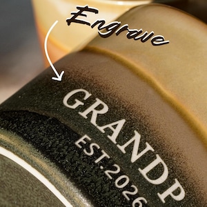 May include: Close-up of a ceramic mug with the word "GRANDPA" and "EST. 2026" in white lettering. The mug is dark green with a textured surface. The top of a second mug is visible in the background, with the word "Engrave" above it.