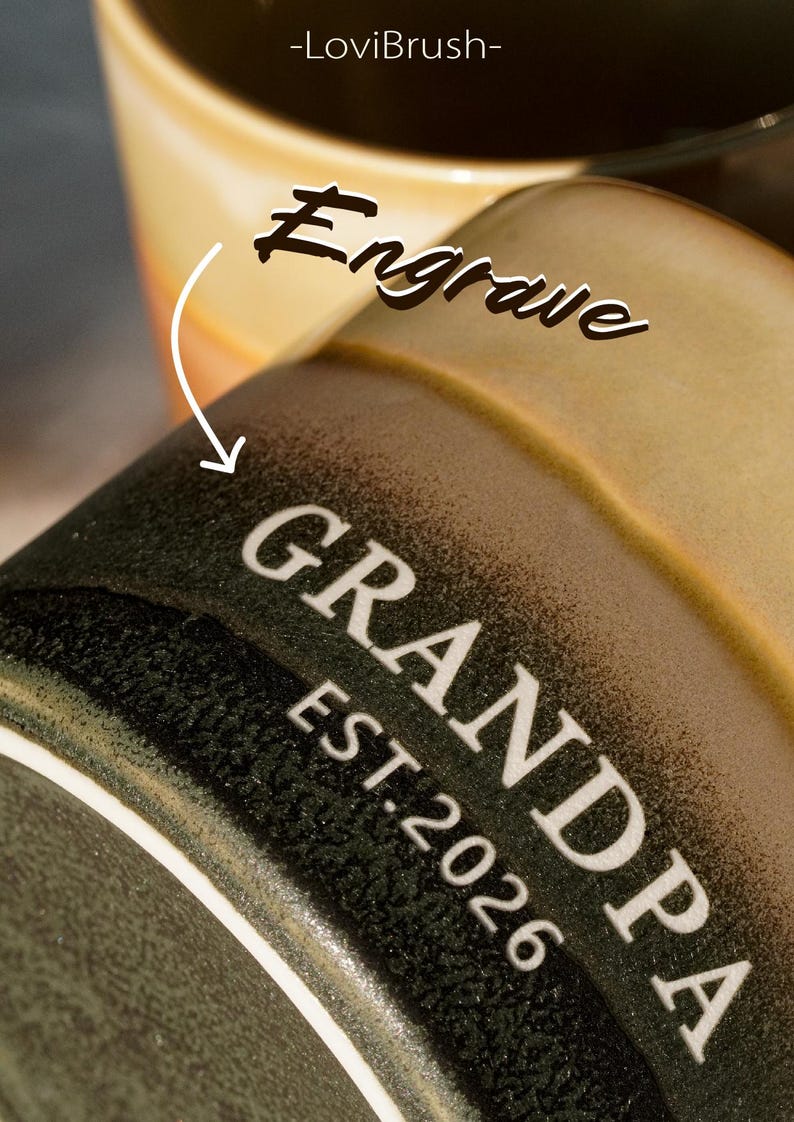 May include: Close-up of a ceramic mug with the word "GRANDPA" and "EST. 2026" in white lettering. The mug is dark green with a textured surface. The top of a second mug is visible in the background, with the word "Engrave" above it.