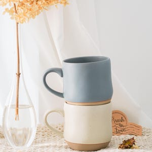 May include: Two ceramic mugs, one on top of the other. The top mug is a muted blue, the bottom is cream-coloured. Both have a brown base and a curved handle. A vase with dried flowers and a wooden sign with "Brush in Love" are in the background.