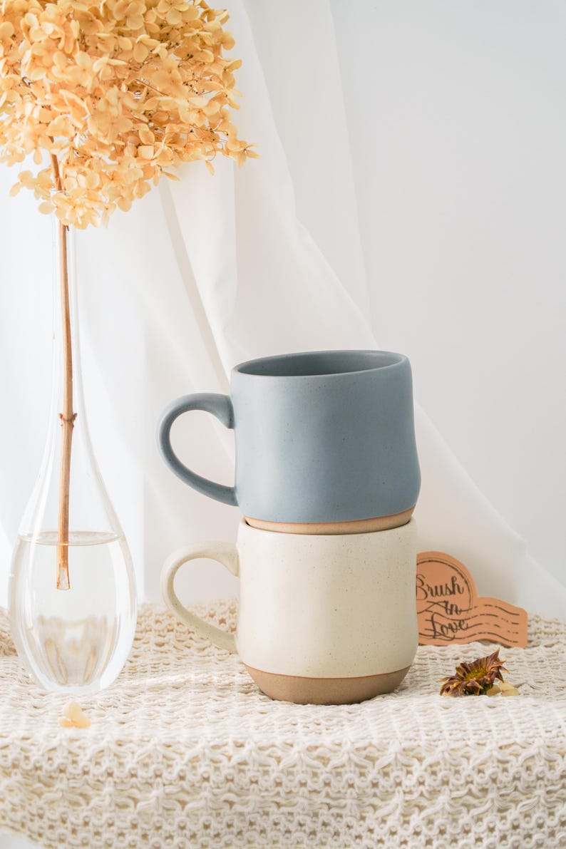 May include: Two ceramic mugs, one on top of the other. The top mug is a muted blue, the bottom is cream-coloured. Both have a brown base and a curved handle. A vase with dried flowers and a wooden sign with "Brush in Love" are in the background.