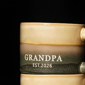 May include: A ceramic mug with a gradient design in shades of beige, brown, and green. The word "GRANDPA" and the year "EST.2026" are printed in white on the mug. The mug has a handle and is likely used for beverages.