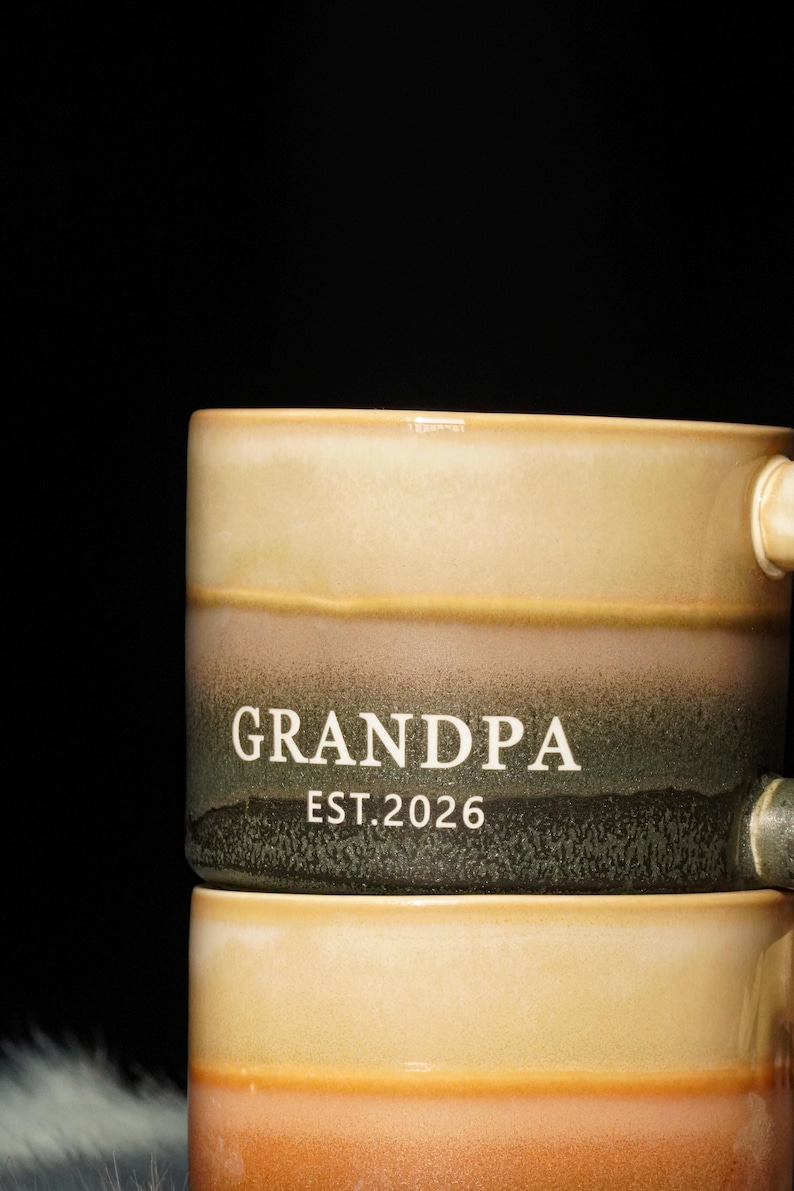May include: A ceramic mug with a gradient design in shades of beige, brown, and green. The word "GRANDPA" and the year "EST.2026" are printed in white on the mug. The mug has a handle and is likely used for beverages.