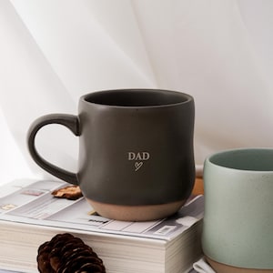 Op de afbeelding: Een donkergrijze keramische mok met het woord "DAD" en een hartsymbool. De mok heeft een afgeronde vorm, een oor en een bruinachtige basis. Hij staat op een boek naast een groene mok.