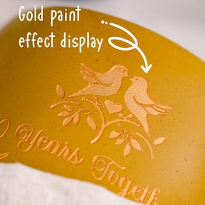 May include: Close-up of a gold-coloured surface with an embossed design of two birds on a branch, with a heart. The text "2 Years Together" is also embossed. The text "Gold paint effect display" is written in white.