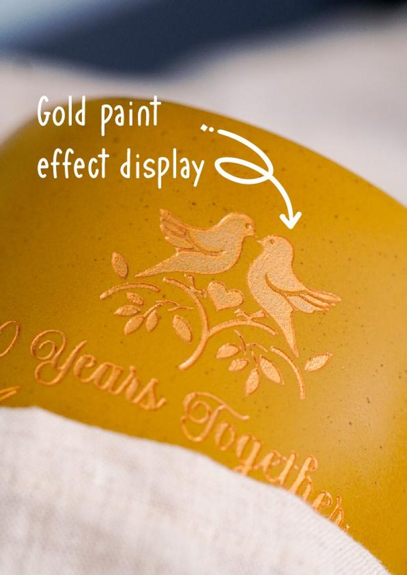 May include: Close-up of a gold-coloured surface with an embossed design of two birds on a branch, with a heart. The text "2 Years Together" is also embossed. The text "Gold paint effect display" is written in white.