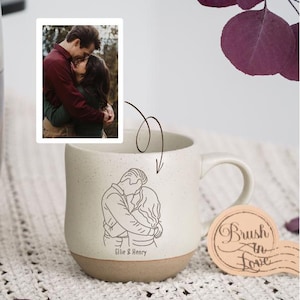 Puede incluir: Taza de cerámica color crema con un dibujo lineal de una pareja abrazándose, con la etiqueta "Ellie & Henry". Una pequeña foto de la pareja está junto a la taza. También hay una pegatina con "Brush in Love". La taza tiene una base marrón.