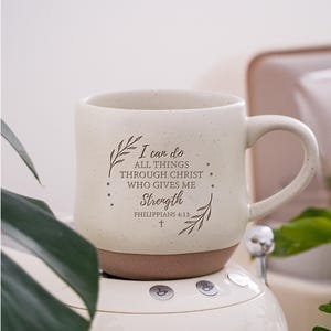 May include: A cream-coloured ceramic mug with a speckled finish and a brown base. The front features dark brown text that reads: I can do ALL THINGS THROUGH CHRIST WHO GIVES ME Strength PHILIPPIANS 4:13, accented by two leafy branches and a small cross.