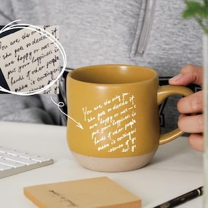 Könnte beinhalten: Senfgelbe Keramik-Tasse mit beigem Boden. Auf der Tasse steht ein Zitat in weißer Schrift: „Du bist die einzige Person, die entscheidet, ob du glücklich bist oder nicht...“ Eine Tastatur und ein Notizblock sind im Hintergrund.