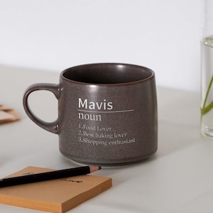 May include: A dark gray ceramic mug with a white inscription that reads "Mavis noun 1. Food Lover 2. Best baking lover 3. Shopping enthusiast".