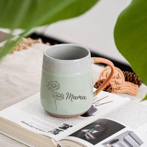 May include: A ceramic mug with a light green base and a white top section. The mug features a floral design and the word "Mama" in script. The handle is a peach color. The mug is sitting on an open book.