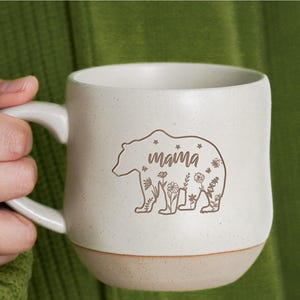 May include: White ceramic mug with a brown bear silhouette and the word "mama" inside the bear. The bear is surrounded by small floral designs.