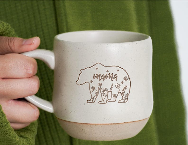 May include: White ceramic mug with a brown bear silhouette and the word "mama" inside the bear. The bear is surrounded by small floral designs.