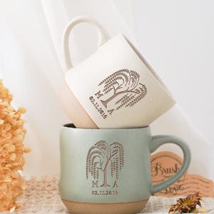 May include: Two ceramic mugs stacked on top of each other. The top mug is off-white with a light brown base, and the bottom mug is a muted green with a light brown base. Both mugs feature a tree design and the date 02.11.2016.